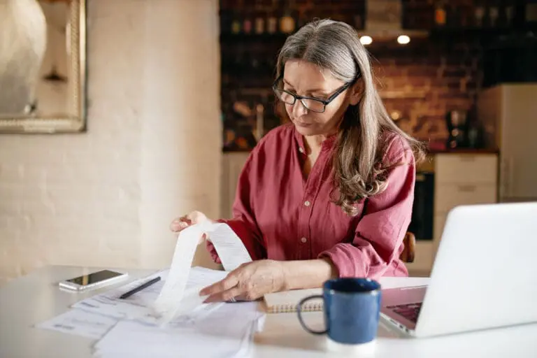 Serious focused middle aged female accountant in eyewear working from home providing outsource bookkeeping service, sitting at table with portable computer, calculating finances, doing paperwork