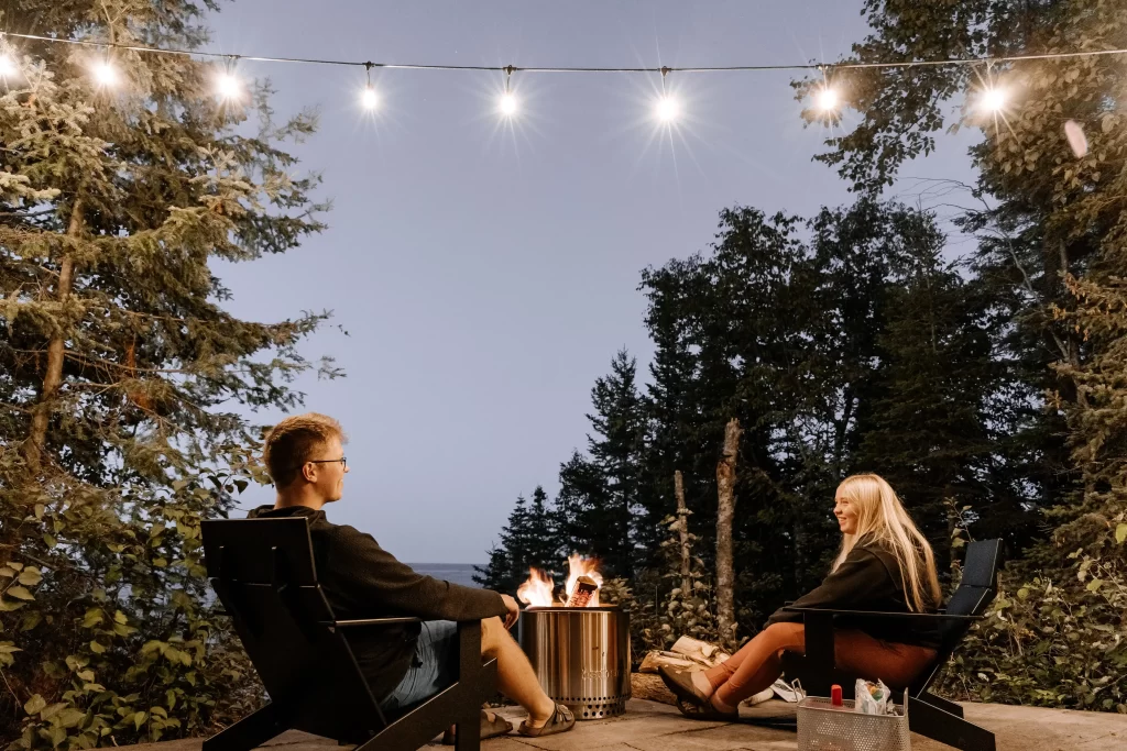 A man in a black jumper and a blonde girl in a black hoodie sit by an outdoor firepit under string lights