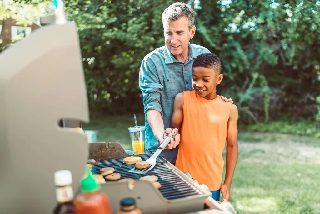 older white man in blue shirt shows young black boy in an orange top how to grill a burger. HOA outdoor amenities.