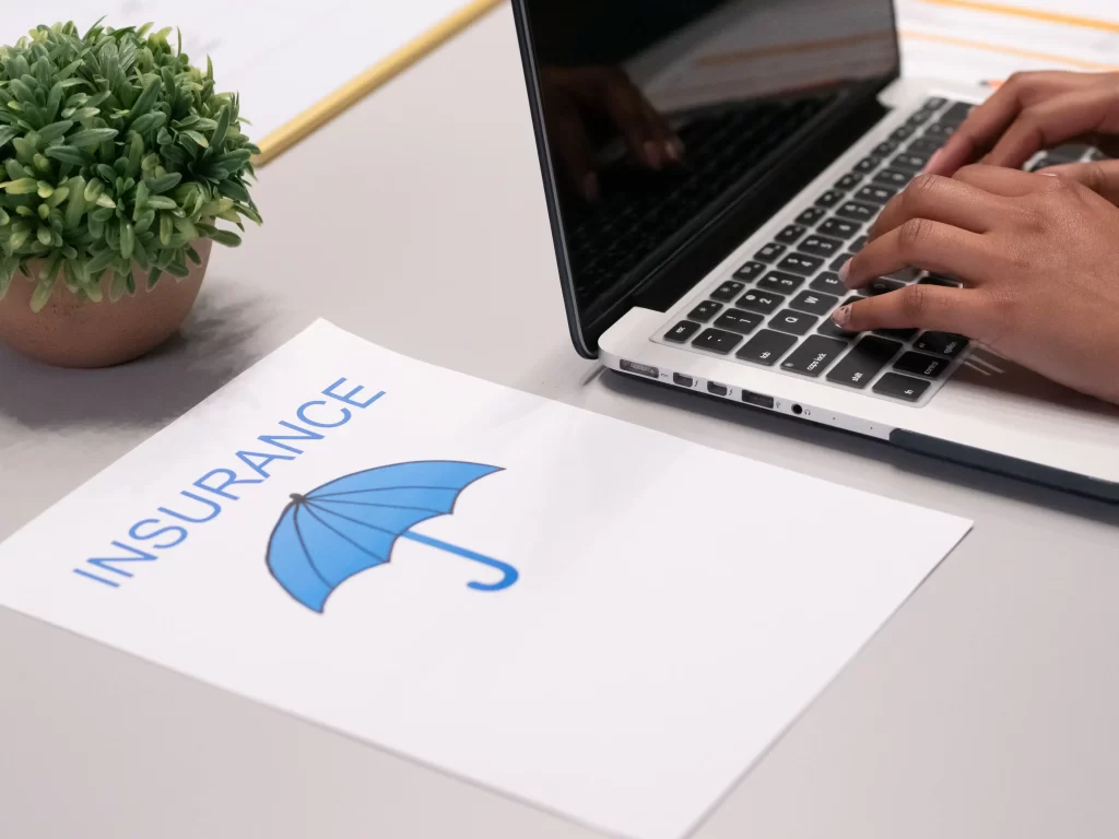 image of a grey desk with a little green plant, a laptop with hands resting on the keyboard and a page with a picture of a blue umbrella and the word insurance. Representing a HOA board member searching for community insurance