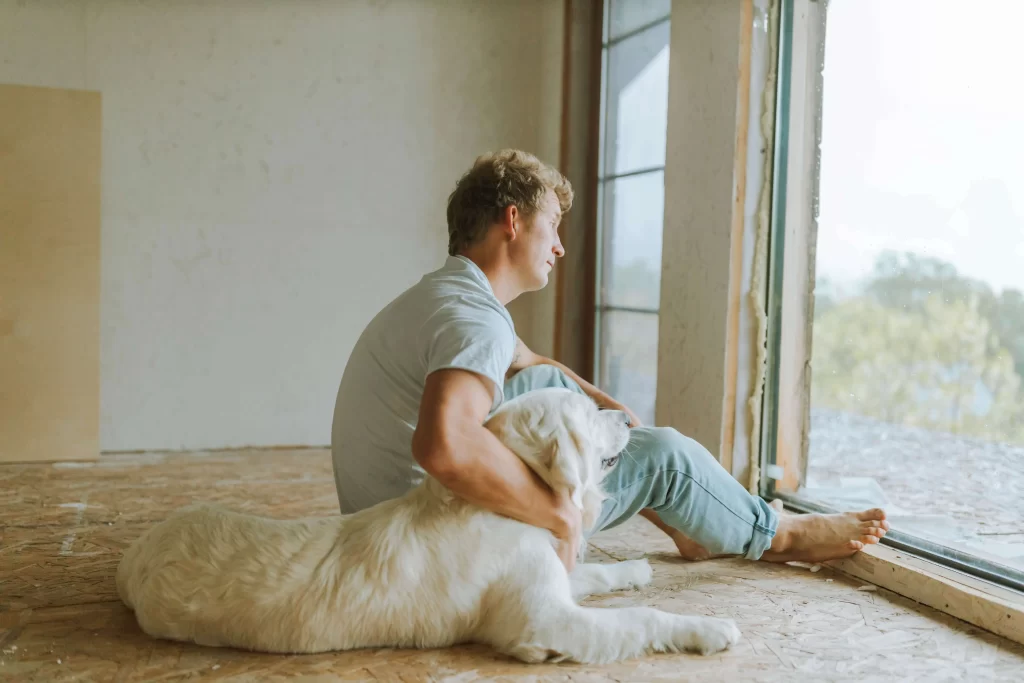 picture of a man with blonde hair looking out of a windom, with his arm around a fluffy white golden retriever