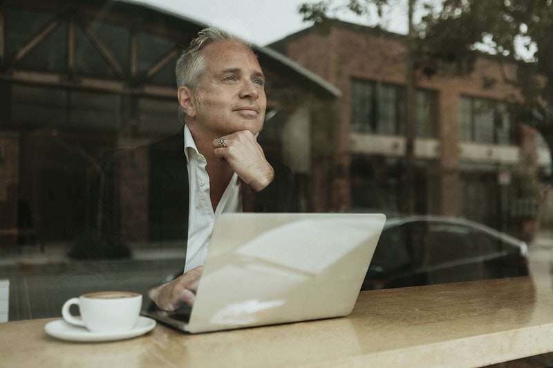 Older man with white hair in a jacket and white shirt looking reflective looking out of the window with a laptop and coffee next to him, thinking about HOA Newsletter