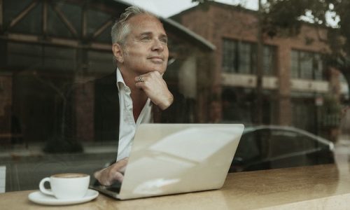 Older man with white hair in a jacket and white shirt looking reflective looking out of the window with a laptop and coffee next to him, thinking about HOA Newsletter