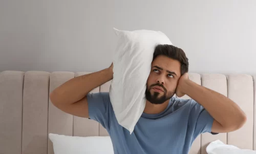 A frustrated man holds a pillow to his head, seeking comfort and contemplating a potential HOA noise complaints due to disruptive sounds in the background.