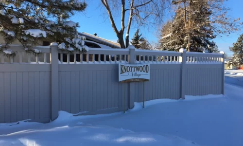 Snowy light brown fence with a sign that says Knottwood Village. There is snow piled up in front and with trees behind