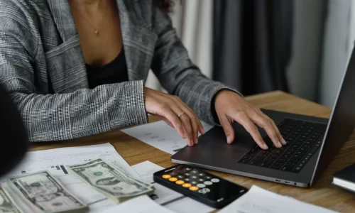image of a woman's torso with a grey jacket, black top and a gold necklace on a laptop with papers, calulculator and money on the desk near her. Represents trying to reduce maintenance costs in her HOA