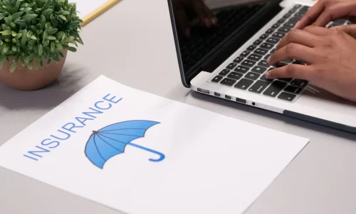 image of a grey desk with a little green plant, a laptop with hands resting on the keyboard and a page with a picture of a blue umbrella and the word insurance. Representing a HOA board member searching for community insurance