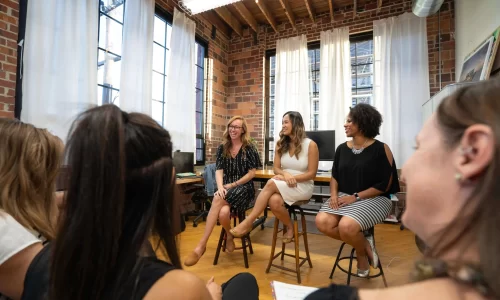 3 women sit on high stools in a brick room with wooden floors and address a group of people (mainly women). Representing a meeting in an efficient HOA