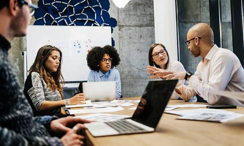 A group of three young women and two men of different ethnicities are in a business meeting in a modern day office. A bald man is talking to the group while there are laptops and documents on the table.